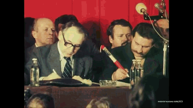 a group of men are sitting at a table and one of them is holding a red pen