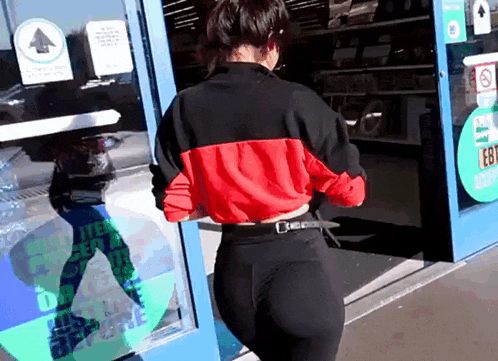 a woman in a red and black jacket and black leggings is walking through a store door .