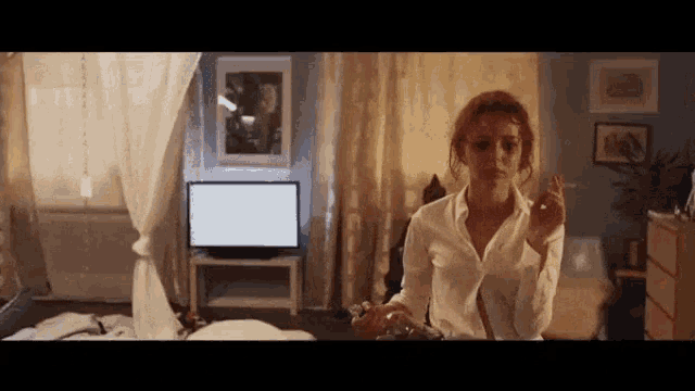 a woman in a white shirt is sitting on a bed smoking a cigarette in front of a television .