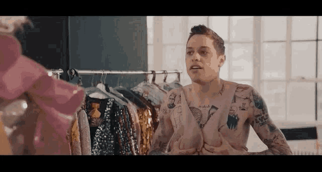 a shirtless man with tattoos is standing in front of a row of clothes .