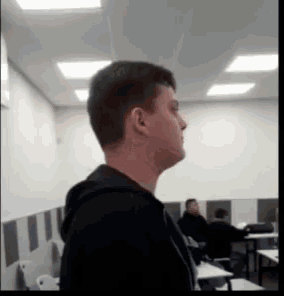 a man in a black hoodie is standing in a classroom with people sitting at tables .