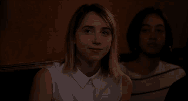 a woman in a white shirt looks at the camera while another woman sits behind her