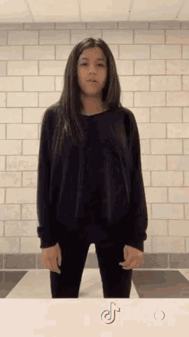 a girl in a black shirt is standing in front of a brick wall with a tiktok logo behind her