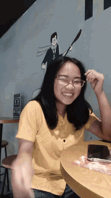 a girl wearing glasses and a yellow shirt is sitting at a table and smiling