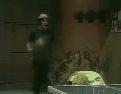 a man in a hat is standing next to a man playing ping pong