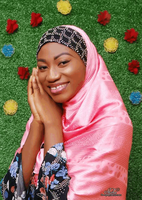 a woman wearing a pink scarf and a black headband is smiling in front of some flowers