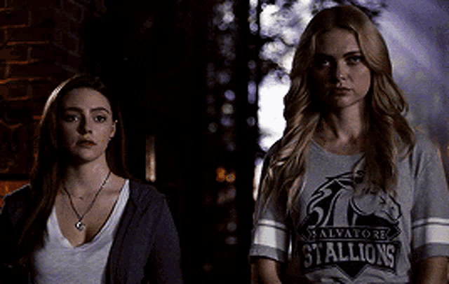 two women are standing next to each other in a dark room . one of the women is wearing a shirt that says stallions .