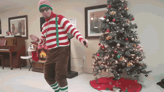 a man in a striped sweater and suspenders is dancing in front of a christmas tree