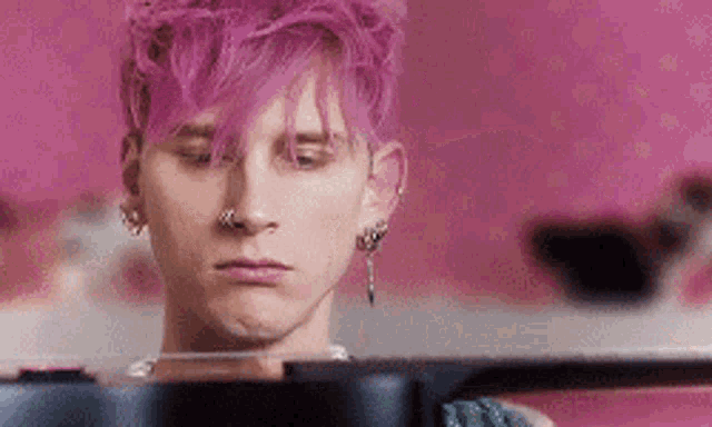 a man with purple hair is sitting in front of a pink wall and looking at something .