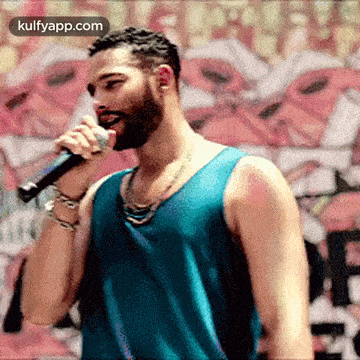 a man in a blue tank top is singing into a microphone in front of a graffiti wall .