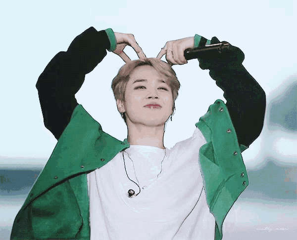 a young man wearing a green jacket and a white shirt making a heart shape with his hands