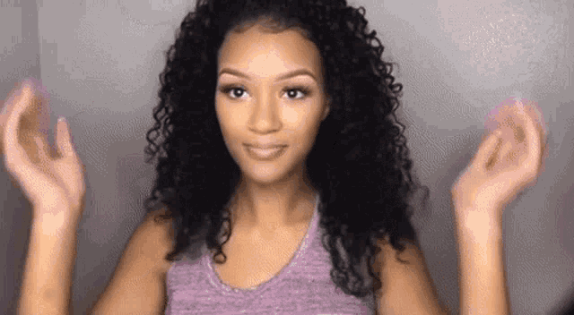 a woman with curly hair is wearing a purple tank top and making a peace sign with her hands .