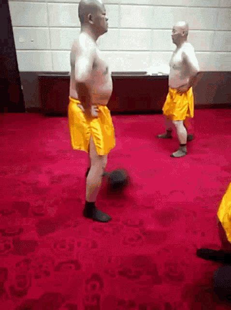 two bald men in yellow shorts are standing on a red rug