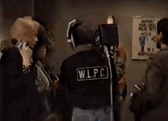 a man wearing a w.l.p.c. vest talks to a man in a suit