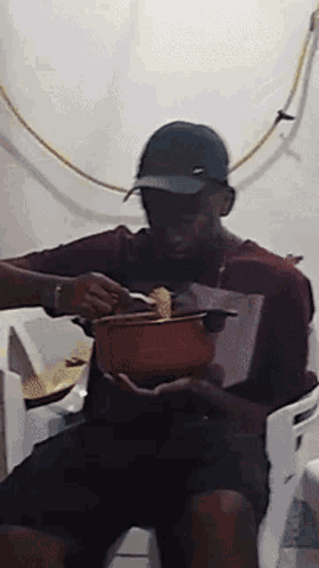 a man wearing a hat is sitting in a chair eating from a pot