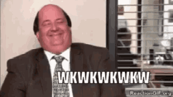 a bald man in a suit and tie is smiling and laughing while sitting in front of a window .