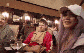 a woman with purple hair is sitting at a table with a man in a red jacket and a woman in a white hat .