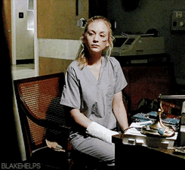 a woman in scrubs is sitting at a desk with blakehelps written on the bottom right