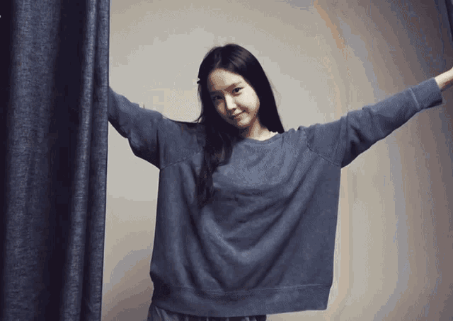 a woman in a blue sweater stands in front of a curtain with her arms outstretched