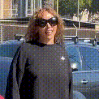a woman wearing sunglasses and a black sweater is standing in front of a parking lot .