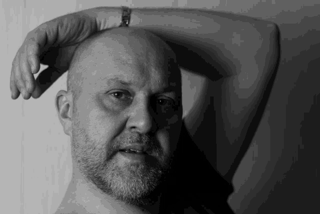 a bald man with a beard holds his hand to his forehead