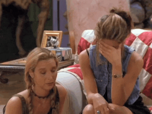 two women are sitting on a couch one is covering her face with her hand .