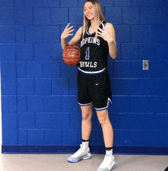 a female basketball player for the hopkins bulls holds a basketball in front of a blue wall