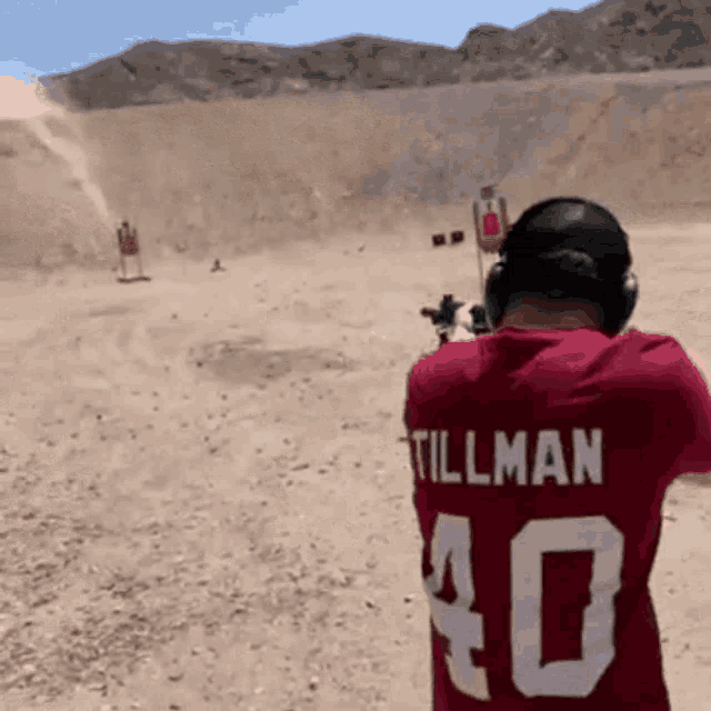 a man wearing a red shirt with tillman on the back is shooting a gun