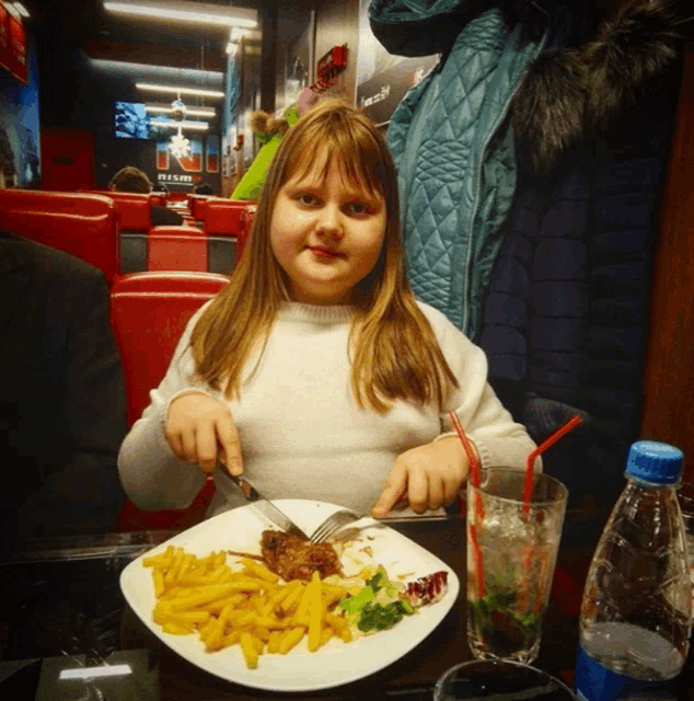 a little girl is sitting at a table with a plate of food and a drink