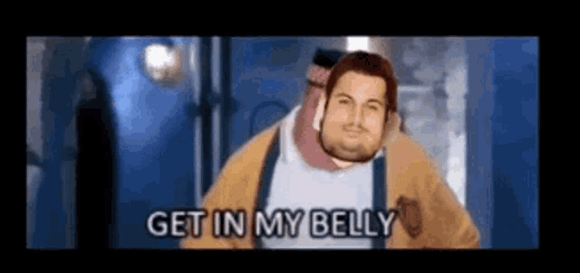 a man with a beard is standing in front of a blue wall and says get in my belly