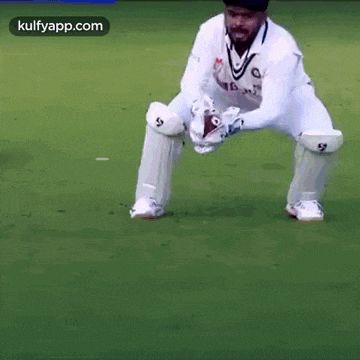 a man in a white cricket uniform is kneeling on the field .