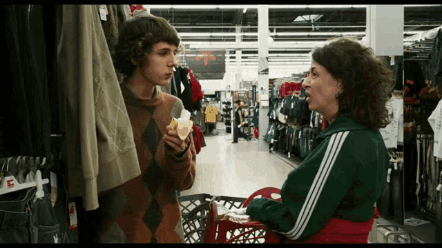 a woman is pushing a shopping cart while a boy holds a sandwich in a clothing store