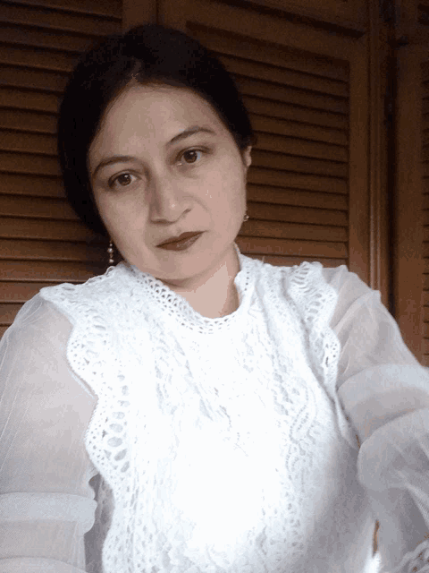 a woman wearing a white lace top stands in front of a wooden shutter