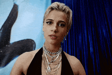 a woman wearing a black top and a necklace is standing in front of a blue curtain .