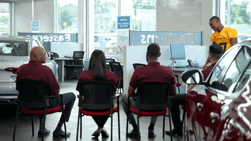 a man in a yellow shirt with the number 7 on it talks to a group of people in a car dealership