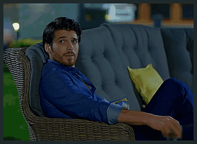 a man in a blue shirt sits in a wicker chair with his legs crossed