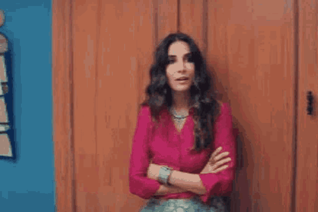 a woman in a pink shirt and green skirt is standing with her arms crossed in front of a wooden wall .