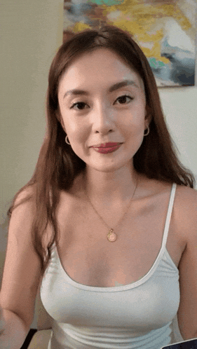 a woman wearing a white tank top and a necklace looks at the camera