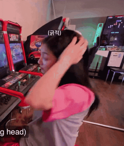 a woman adjusts her hair while playing a video game in an arcade with the words ' g head ' below her