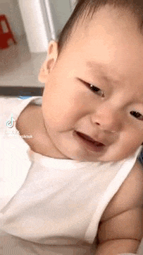 a baby in a white tank top is crying while looking at the camera .