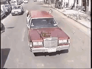a red cadillac is driving down a street with a dog on the hood .