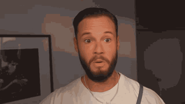 a man with a beard wearing a white t-shirt and a necklace with the letter m on it