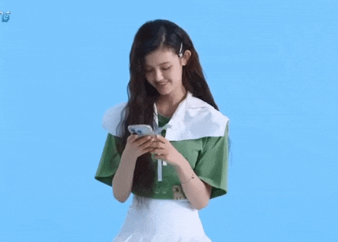 a girl in a green shirt and white skirt is holding a cell phone in her hands .