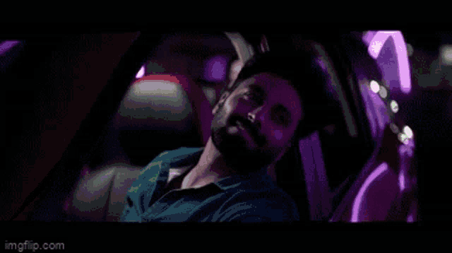 a man with a beard is sitting in a car with green lights behind him .