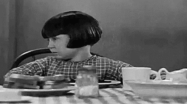 a black and white photo of a young girl sitting at a table with plates of food .