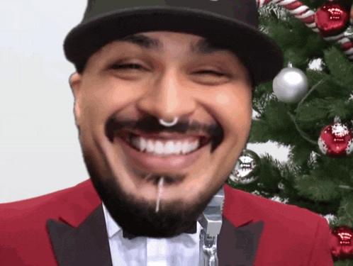 a man with a beard wearing a hat and a red jacket is smiling in front of a christmas tree