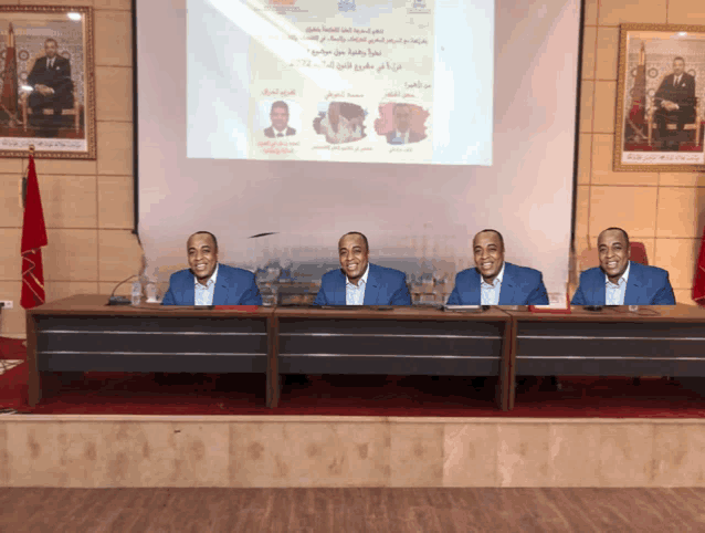 four men sit at a table in front of a projector screen with arabic writing
