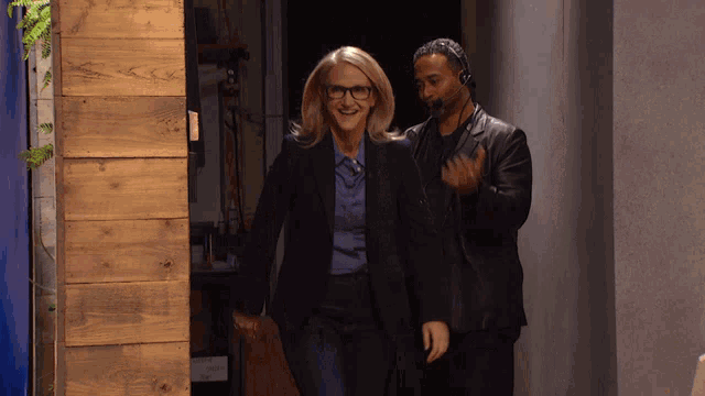 a woman in glasses and a man in a leather jacket are walking down a hallway