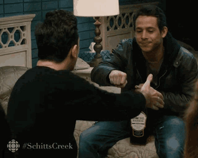 two men sitting on a bed with a bottle of bourbon