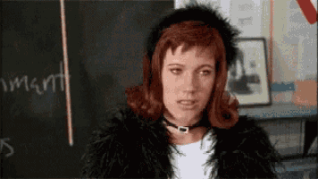 a woman with red hair and a black fur coat is standing in front of a blackboard in a classroom .